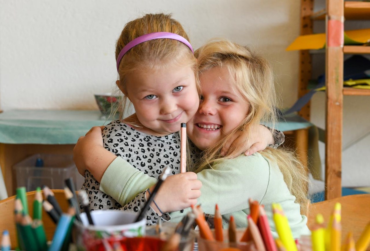Zwei kleine Mädchen im Kindergarten umarmen sich herzlich und blicken lächelnd in die Kamera. Ein Mädchen hat einen Stift in der Hand.